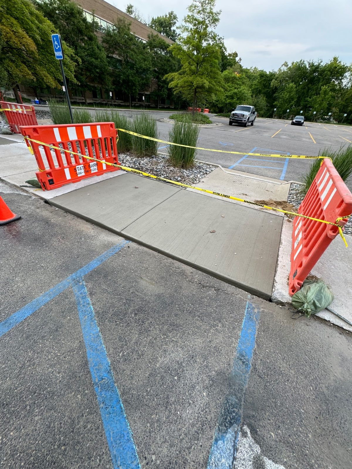 Newly poured broom-finished sidewalk panel and accessibility ramp at a commercial parking lot, protected by orange safety barricades