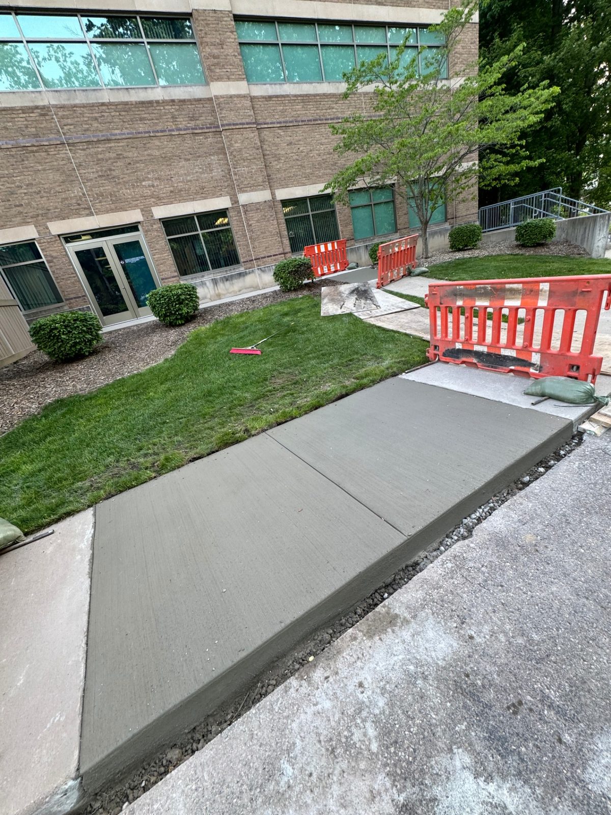 Newly replaced concrete sidewalk panels outside a commercial office building with safety barricades and open base trenches alongside
