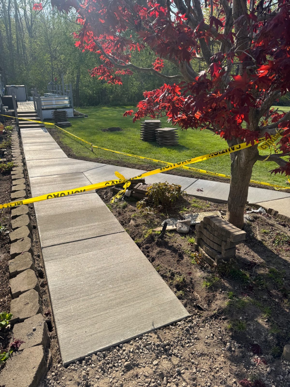New concrete backyard walkway splitting into two branches around a Japanese maple toward deck steps, with caution tape and forms in place