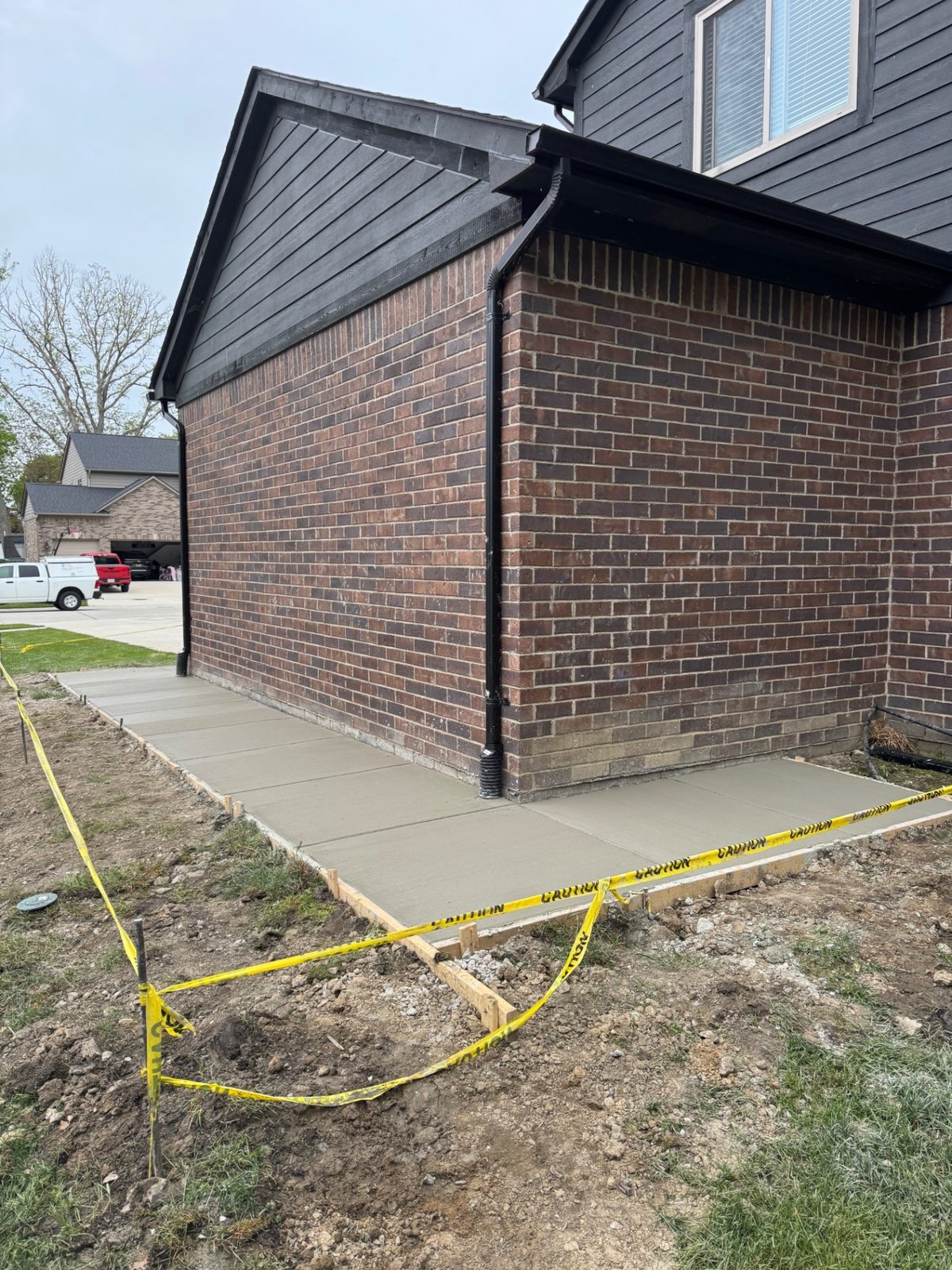 New concrete walkway wrapping the corner of a two-story brick home with wooden forms and caution tape still in place